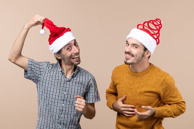 Vista frontal de dos chicos riendo con gorro de Papá Noel, uno mostrando su sombrero sobre fondo beige aislado