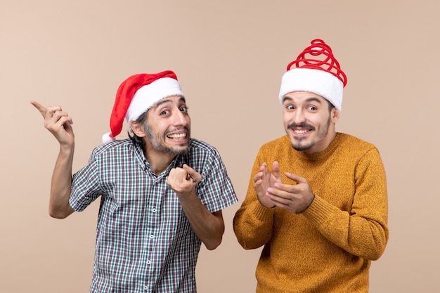 Vista frontal de dos chicos sonrientes con gorro de Papá Noel que muestra algo sobre fondo beige aislado