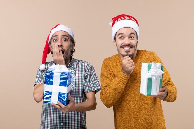 Vista frontal de dos chicos un sorprendido y feliz vistiendo gorro de Papá Noel y sosteniendo sus regalos sobre fondo beige aislado