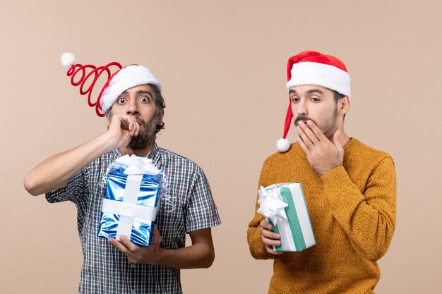 Vista frontal de dos chicos sorprendidos con gorro de Papá Noel y poniendo sus manos en la boca sobre fondo beige aislado