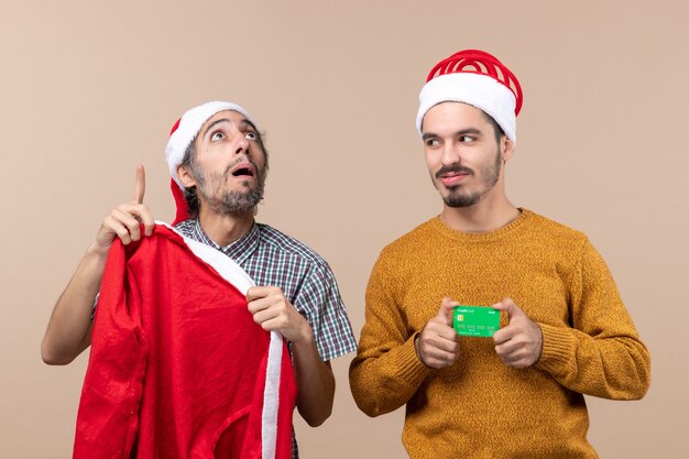 Foto gratuita vista frontal de dos chicos, uno sosteniendo el abrigo de santa y mirando alto y el otro con tarjeta de crédito mirando a su amigo sobre fondo beige aislado