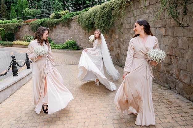 Vista frontal de dos damas de honor en vestidos largos de noche que sostienen arreglos florales y miran a la novia que usa un vestido de novia de moda y se gira mientras camina por la calle antigua