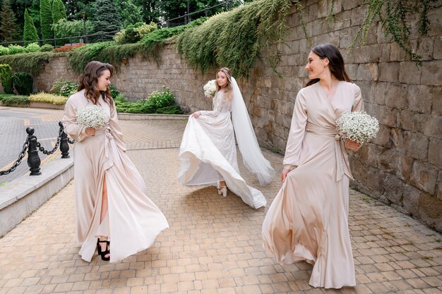 Vista frontal de dos damas de honor en vestidos largos de noche que sostienen arreglos florales y miran a la novia que usa un vestido de novia de moda y se gira mientras camina por la calle antigua