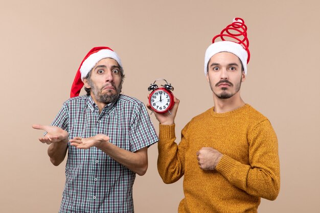 Vista frontal de dos hombres confundidos uno sosteniendo un reloj de alarma sobre fondo beige aislado