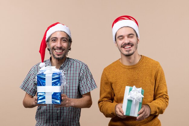 Vista frontal de dos hombres felices con gorro de Papá Noel y sosteniendo regalos de Navidad sobre fondo beige aislado