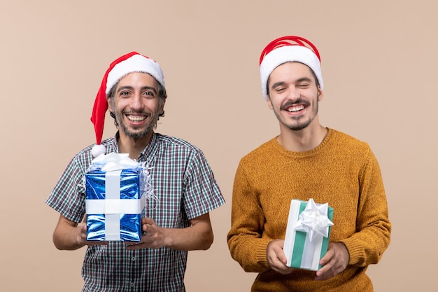 Vista frontal de dos hombres felices con gorro de Papá Noel y sosteniendo regalos de Navidad sobre fondo beige aislado