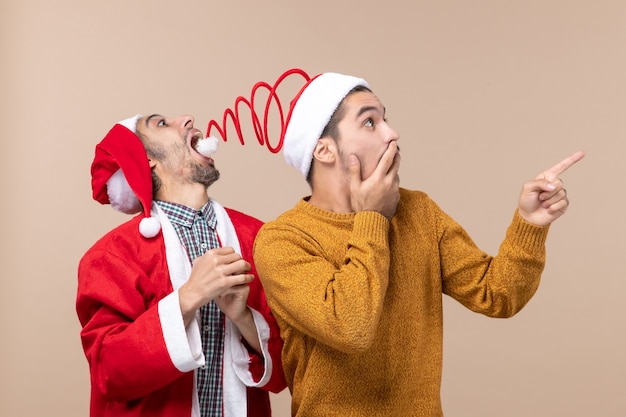 Foto gratuita vista frontal de dos hombres con gorro de papá noel, uno tratando de atrapar sombreros pompón y el otro mostrando la dirección sobre fondo beige aislado