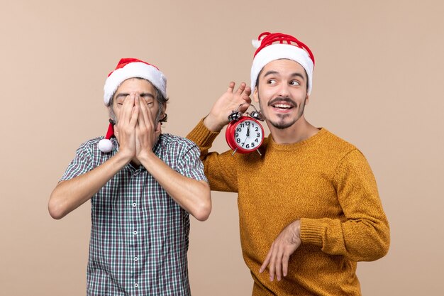 Vista frontal de dos hombres de Navidad, uno cubriendo la cara con las manos y el otro sosteniendo un reloj de alarma sobre fondo beige aislado
