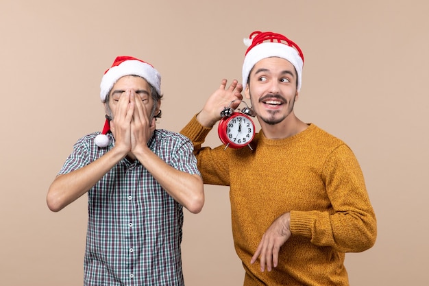 Vista frontal de dos hombres de Navidad, uno cubriendo la cara con las manos y el otro sosteniendo un reloj de alarma sobre fondo beige aislado