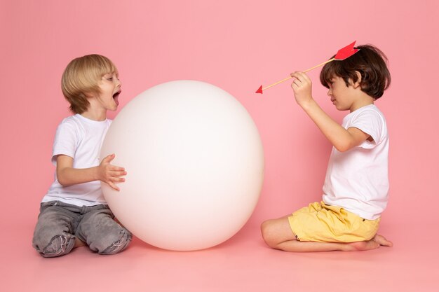 Una vista frontal de dos niños jugando entre sí a través de una pelota redonda blanca con camisetas blancas en el piso rosa