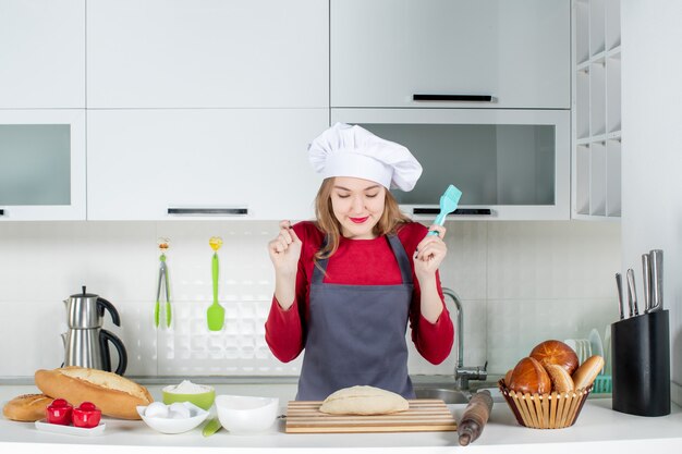 Vista frontal dulce mujer rubia con sombrero de cocinero y delantal mirando pan en la cocina