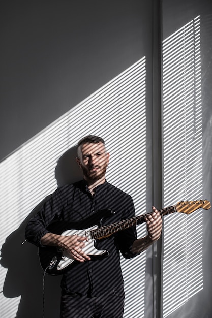 Vista frontal del ejecutante masculino tocando la guitarra eléctrica junto a la ventana