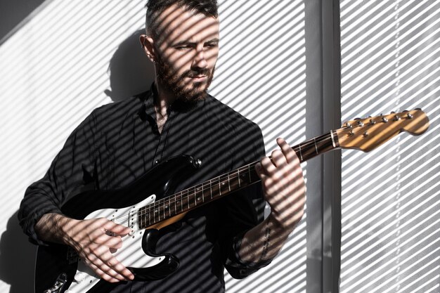 Vista frontal del ejecutante masculino tocando la guitarra eléctrica con sombras de persianas