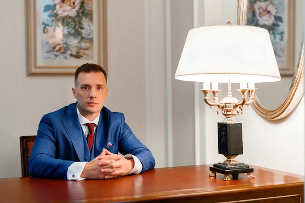 Vista frontal de un elegante hombre de negocios con traje de moda que mantiene las manos juntas mirando seriamente a la cámara mientras se sienta en una mesa de madera con una lámpara vintage en el fondo de la pared con una foto