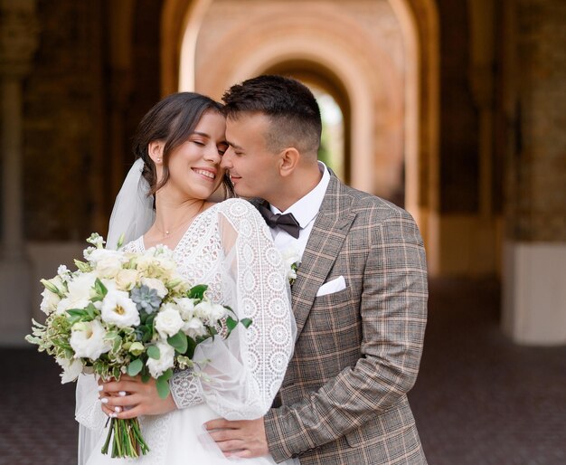 Vista frontal del elegante novio en traje que de pie y abrazando a la novia Mujer feliz en vestido de novia blanco sosteniendo hermosa