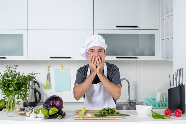 Vista frontal emocionado guapo cocinero de sexo masculino en uniforme de pie detrás de la mesa de la cocina