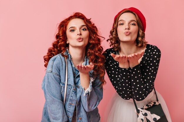 Vista frontal de encantadoras damas enviando besos al aire. Foto de estudio de mujeres bonitas expresando amor sobre fondo rosa.