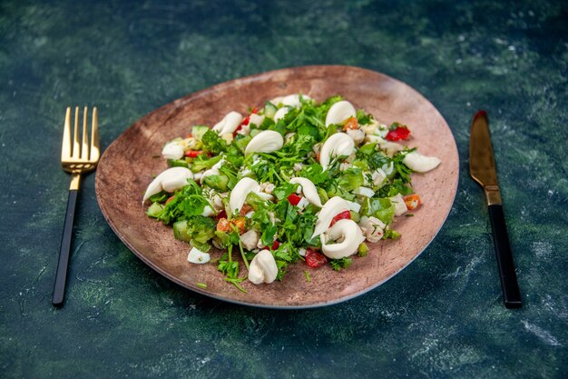 Vista frontal ensalada de verduras frescas dentro de un elegante plato con cubiertos sobre fondo azul oscuro