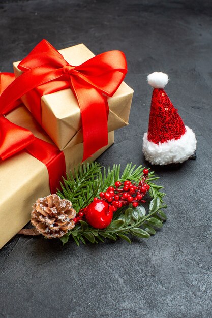 Vista frontal del estado de ánimo navideño con hermosos regalos con cinta en forma de arco y accesorios de decoración de ramas de abeto sombrero de santa claus conos de coníferas sobre un fondo oscuro