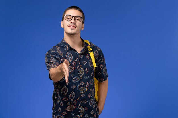 Vista frontal del estudiante en camisa de algodón oscuro con mochila amarilla un apretón de manos en la pared azul