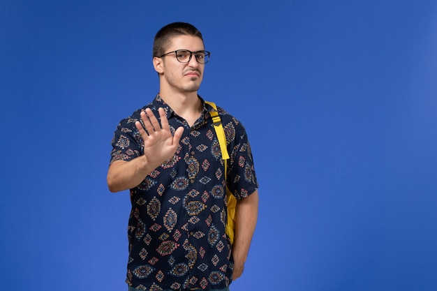 Vista frontal del estudiante en camisa de algodón oscuro con mochila amarilla posando en la pared azul