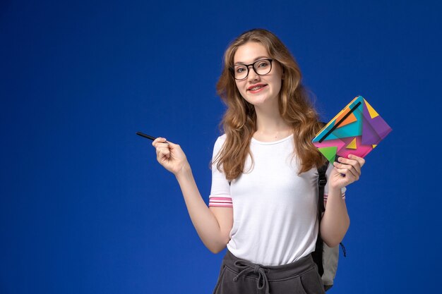 Vista frontal de la estudiante en camisa blanca con lápiz y cuaderno en la pared azul