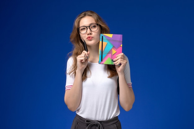 Vista frontal de la estudiante en camisa blanca con lápiz y cuaderno pensando en la pared azul
