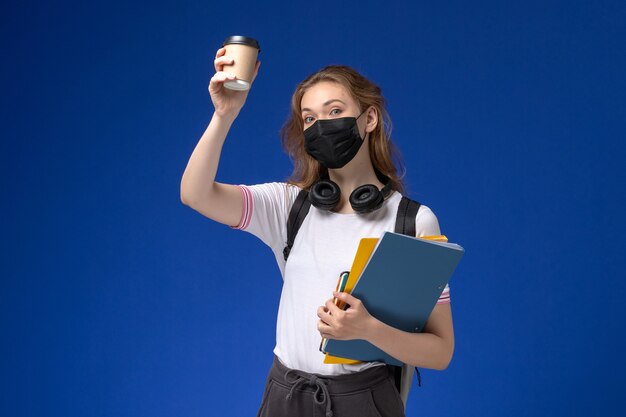 Vista frontal de la estudiante en camisa blanca con mochila máscara negra sosteniendo café y archivos en la pared azul