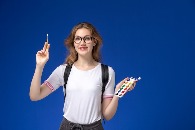 Vista frontal de la estudiante en camisa blanca con mochila y sosteniendo un pincel de pintura artística sonriendo en la pared azul