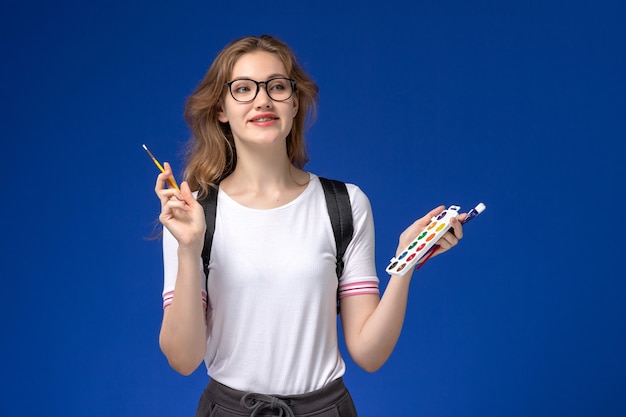 Vista frontal de la estudiante en camisa blanca con mochila y sosteniendo pinceles de pintura de arte en la pared azul