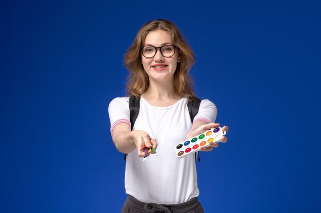 Vista frontal de la estudiante en camisa blanca con mochila y sosteniendo pinceles de pintura de arte en la pared azul