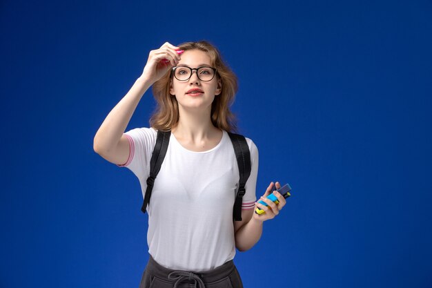 Vista frontal de la estudiante en camisa blanca con mochila y sosteniendo rotuladores en la pared azul