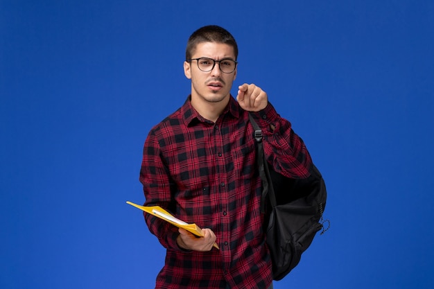 Vista frontal del estudiante en camisa a cuadros roja con mochila con archivos y cuaderno en la pared azul claro