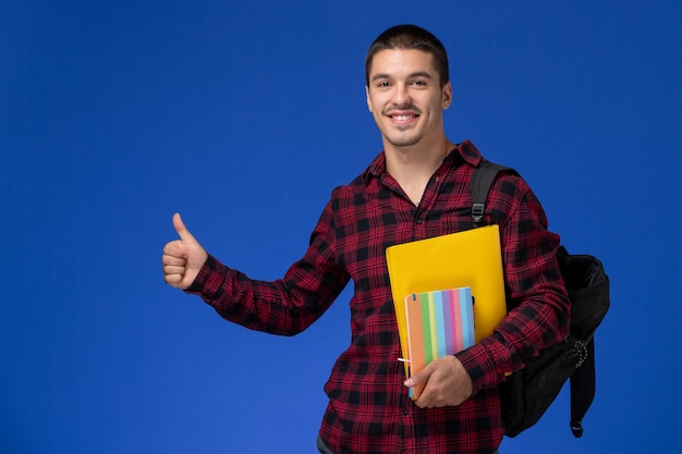 Vista frontal del estudiante en camisa a cuadros roja con mochila con archivos y cuadernos en la pared azul