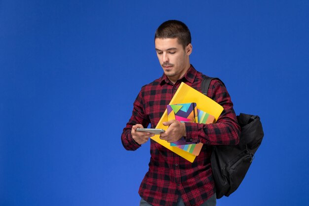 Vista frontal del estudiante en camisa a cuadros roja con mochila con cuaderno y archivos usando su teléfono en la pared azul