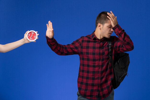 Vista frontal del estudiante en camisa a cuadros roja con mochila posando en la pared azul