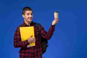 Foto gratuita vista frontal del estudiante en camisa a cuadros roja con mochila sosteniendo archivos amarillos y café en la pared azul