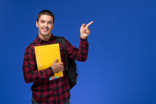 Vista frontal del estudiante en camisa a cuadros roja con mochila sosteniendo archivos amarillos sonriendo en la pared azul