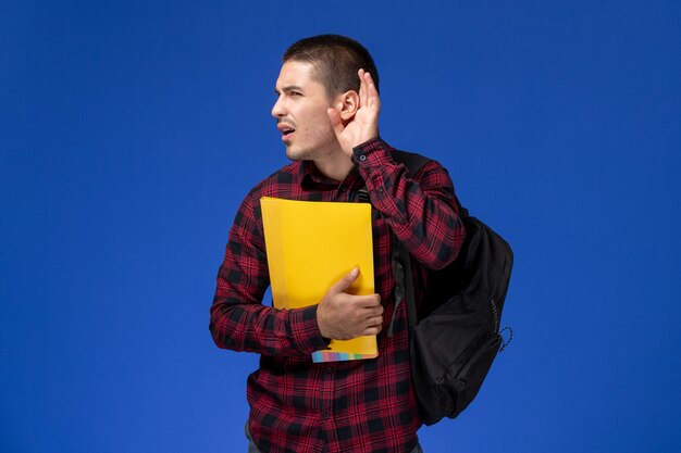 Vista frontal del estudiante en camisa a cuadros roja con mochila sosteniendo archivos amarillos tratando de escuchar en la pared azul