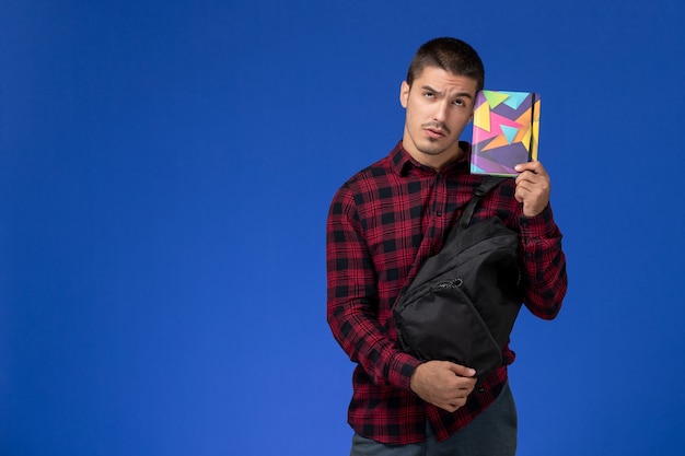 Vista frontal del estudiante en camisa a cuadros roja con mochila sosteniendo cuaderno en la pared azul