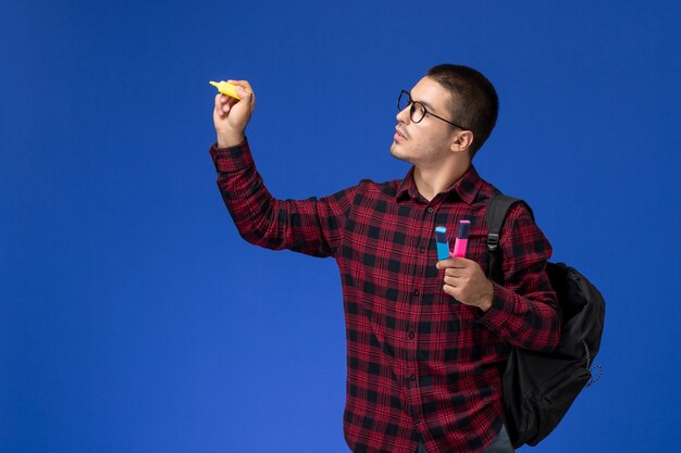 Vista frontal del estudiante en camisa a cuadros roja con mochila sosteniendo rotuladores en la pared azul claro
