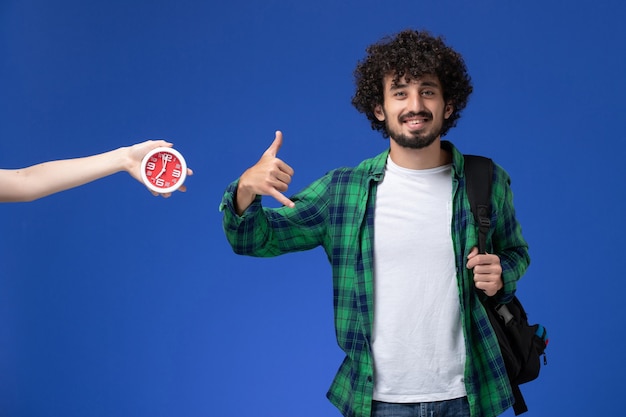 Vista frontal del estudiante en camisa a cuadros verde con mochila negra en el escritorio azul claro