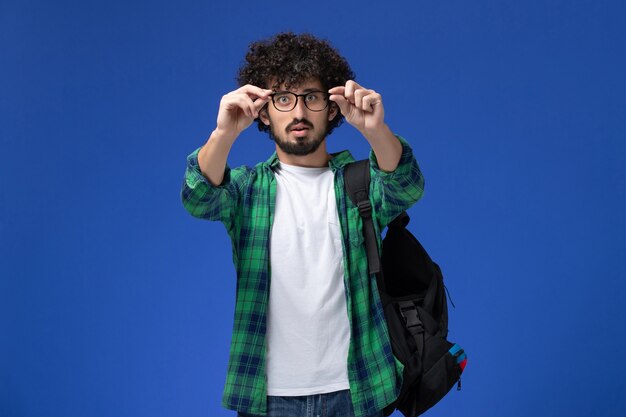Vista frontal del estudiante en camisa a cuadros verde con mochila negra en la pared azul