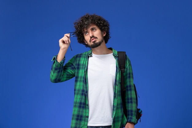 Vista frontal del estudiante en camisa a cuadros verde con mochila negra pensando en la pared azul