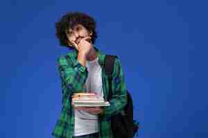 Foto gratuita vista frontal del estudiante en camisa a cuadros verde con mochila negra pensando sosteniendo cuadernos y archivos en la pared azul
