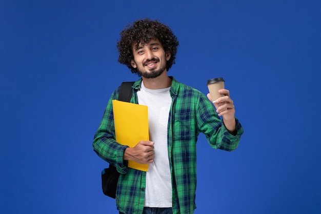 Foto gratuita vista frontal del estudiante en camisa a cuadros verde con mochila negra y sosteniendo archivos y café en la pared azul