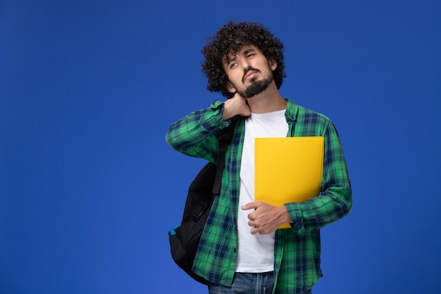 Vista frontal del estudiante en camisa a cuadros verde con mochila negra y sosteniendo archivos con dolor de cuello en la pared azul