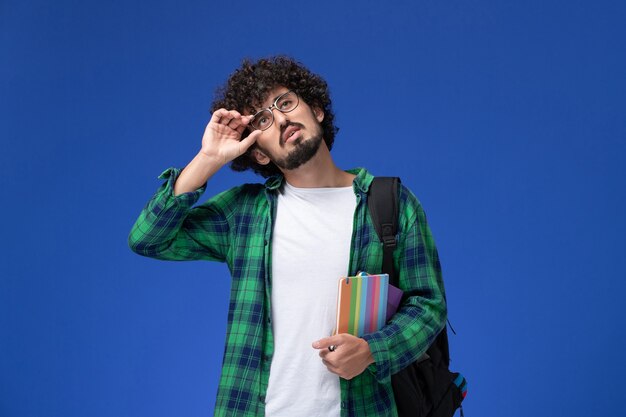 Vista frontal del estudiante en camisa a cuadros verde con mochila negra y sosteniendo el cuaderno en la pared azul