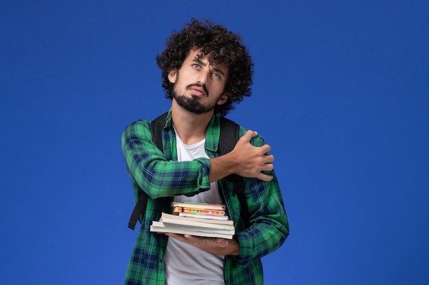 Vista frontal del estudiante en camisa a cuadros verde con mochila negra sosteniendo cuadernos y archivos en la pared azul