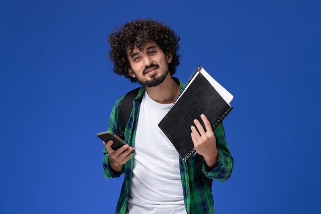 Vista frontal del estudiante en camisa a cuadros verde con mochila negra sosteniendo cuadernos y teléfono en la pared azul